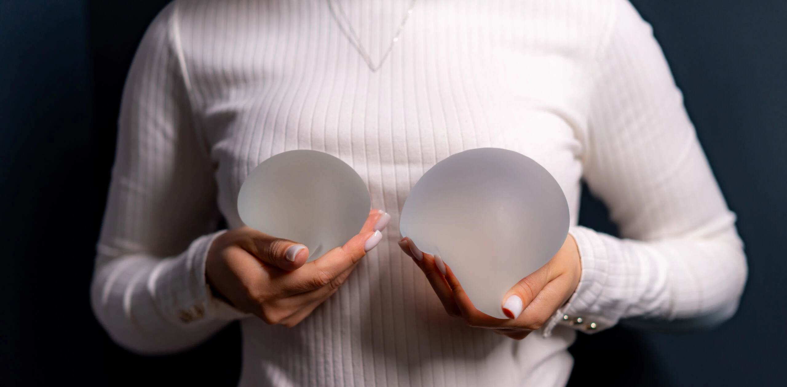 A close-up view of a woman wearing a white long-sleeve ribbed top, holding two different-sized semi-translucent textured silicone breast implants in her hands against a dark background to demonstrate sizing options for a Natural Looking Breast Augmentation in UK.