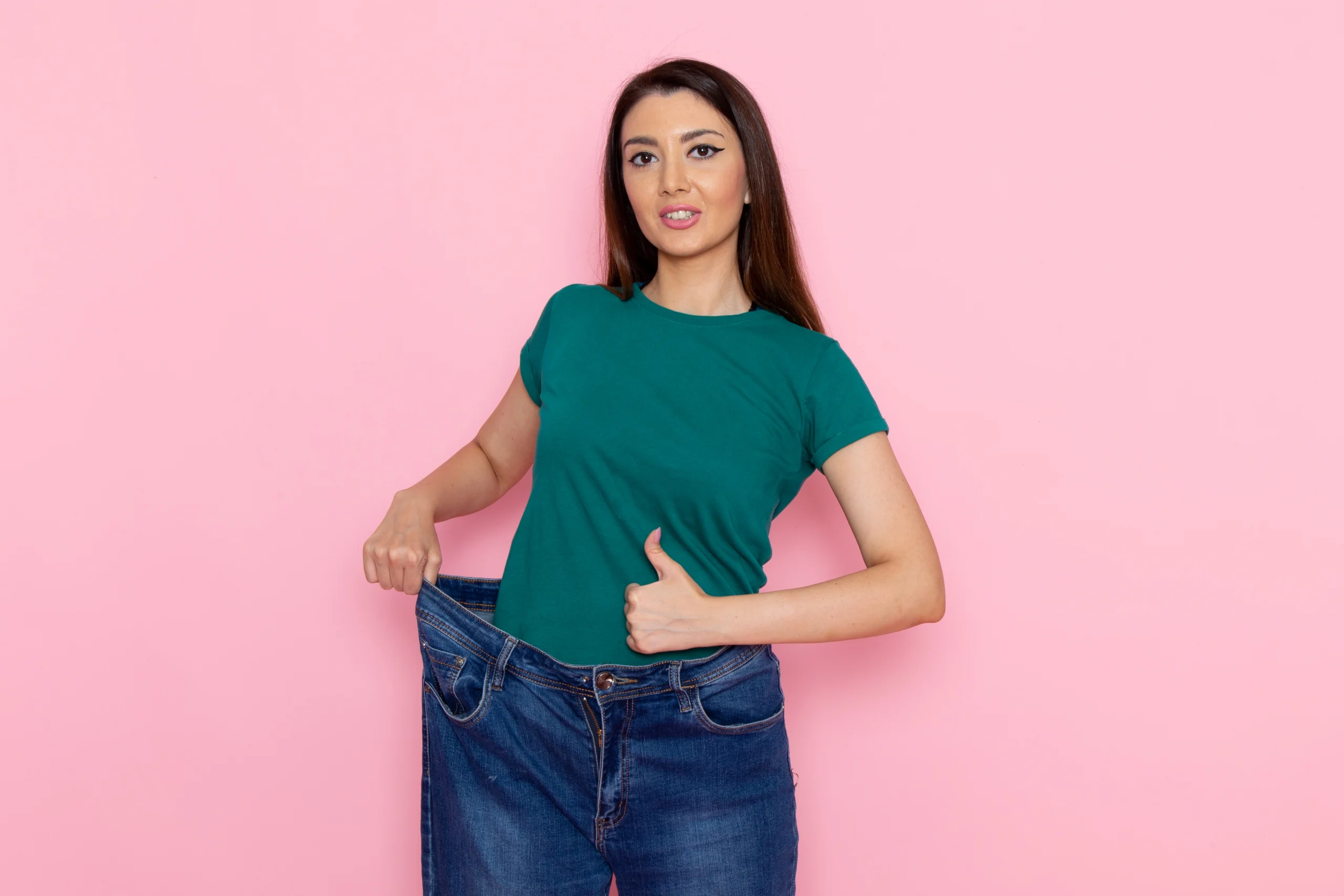 A frontal view of a young woman with her hair in buns, wearing a green cropped t-shirt and grey undergarments against a pink background, using her hands to measure her waist and check if Will my stomach ever be flat after a tummy tuck in UK.