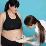 A side-profile view of a woman with dark hair in black activewear standing against a light blue background while a practitioner in a white coat and gloves draws white planning lines across her lower abdomen to map out abdominoplasty safety and recovery in UK.