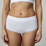 A cropped frontal view of a woman standing in white high-waisted briefs and a crop top, featuring black dashed lines and descending arrows on her waist flanks to map the incision borders for tummy tuck scar healing in UK.