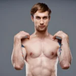 A cropped frontal view of a fit, shirtless man with shoulder tattoos standing against a grey studio backdrop, lifting his hands toward his clavicle area to demonstrate pectoral mobility and map out milestones for Gynecomastia Surgery Recovery in UK.