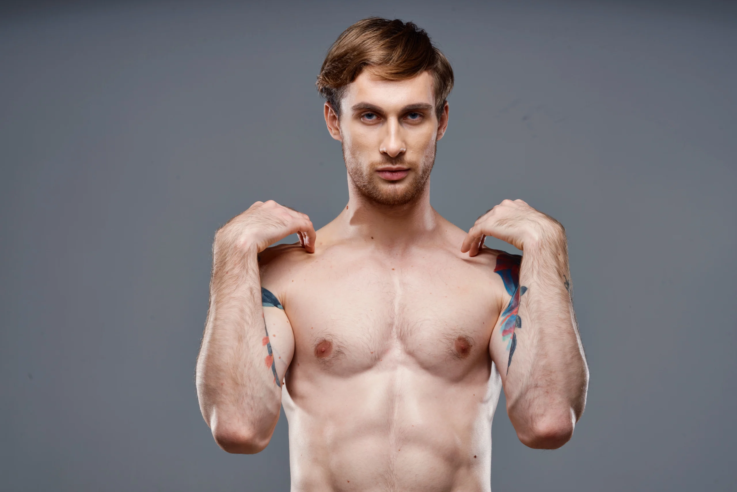 A cropped frontal view of a fit, shirtless man with shoulder tattoos standing against a grey studio backdrop, lifting his hands toward his clavicle area to demonstrate pectoral mobility and map out milestones for Gynecomastia Surgery Recovery in UK.
