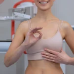 A frontal shot from the neck down of a smiling woman in a beige bralette standing in front of a mammogram machine, making an OK sign with her right hand and gesturing toward her chest to evaluate safety parameters for Breast Augmentation UK.