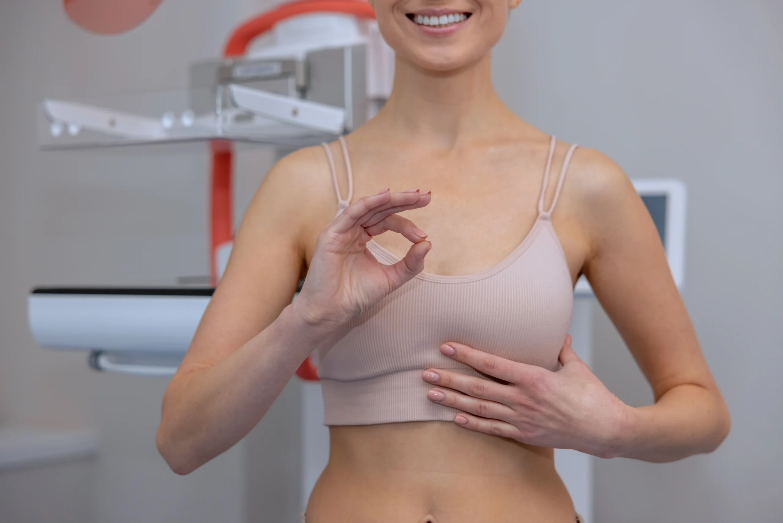 A frontal shot from the neck down of a smiling woman in a beige bralette standing in front of a mammogram machine, making an OK sign with her right hand and gesturing toward her chest to evaluate safety parameters for Breast Augmentation UK.