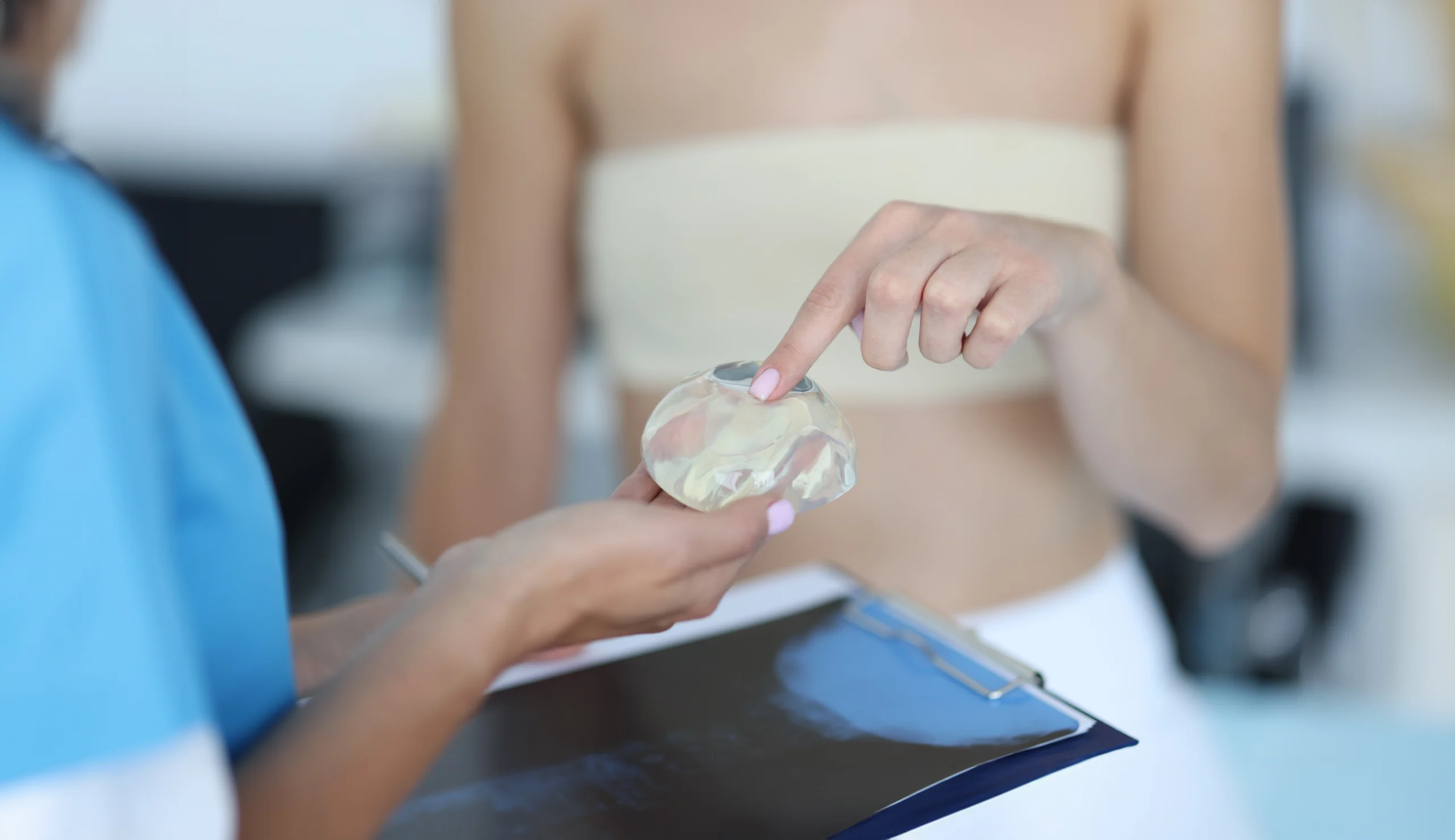An up-close shot of a patient wearing a beige elastic compression bandage across her chest, using her index finger to touch a transparent silicone gel breast implant held by a medical professional to explore options for Hybrid Breast Augmentation UK.