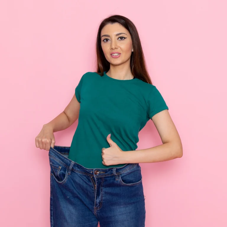 A frontal view of a young woman with her hair in buns, wearing a green cropped t-shirt and grey undergarments against a pink background, using her hands to measure her waist and check if Will my stomach ever be flat after a tummy tuck in UK.