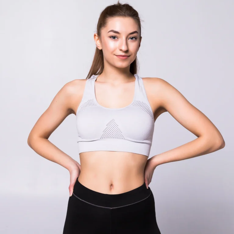 A frontal portrait of a young woman standing with hands on her hips, wearing a light grey sports bra and black leggings against a grey background, displaying a toned, tight lower abdomen to illustrate the Benefit of a Mini Tummy Tuck in UK.