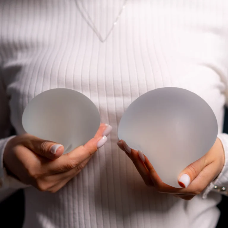 A close-up view of a woman wearing a white long-sleeve ribbed top, holding two different-sized semi-translucent textured silicone breast implants in her hands against a dark background to demonstrate sizing options for a Natural Looking Breast Augmentation in UK.